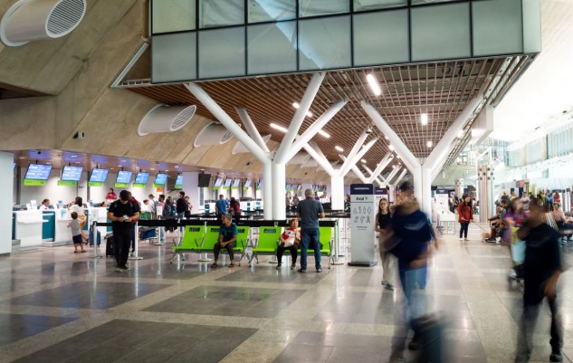 Visão ampla do saguão de check-in de um aeroporto moderno e movimentado. O espaço possui colunas brancas em formato de árvore que sustentam o teto, balcões de atendimento ao fundo com telas azuis e passageiros circulando, alguns representados com efeito de desfoque de movimento.