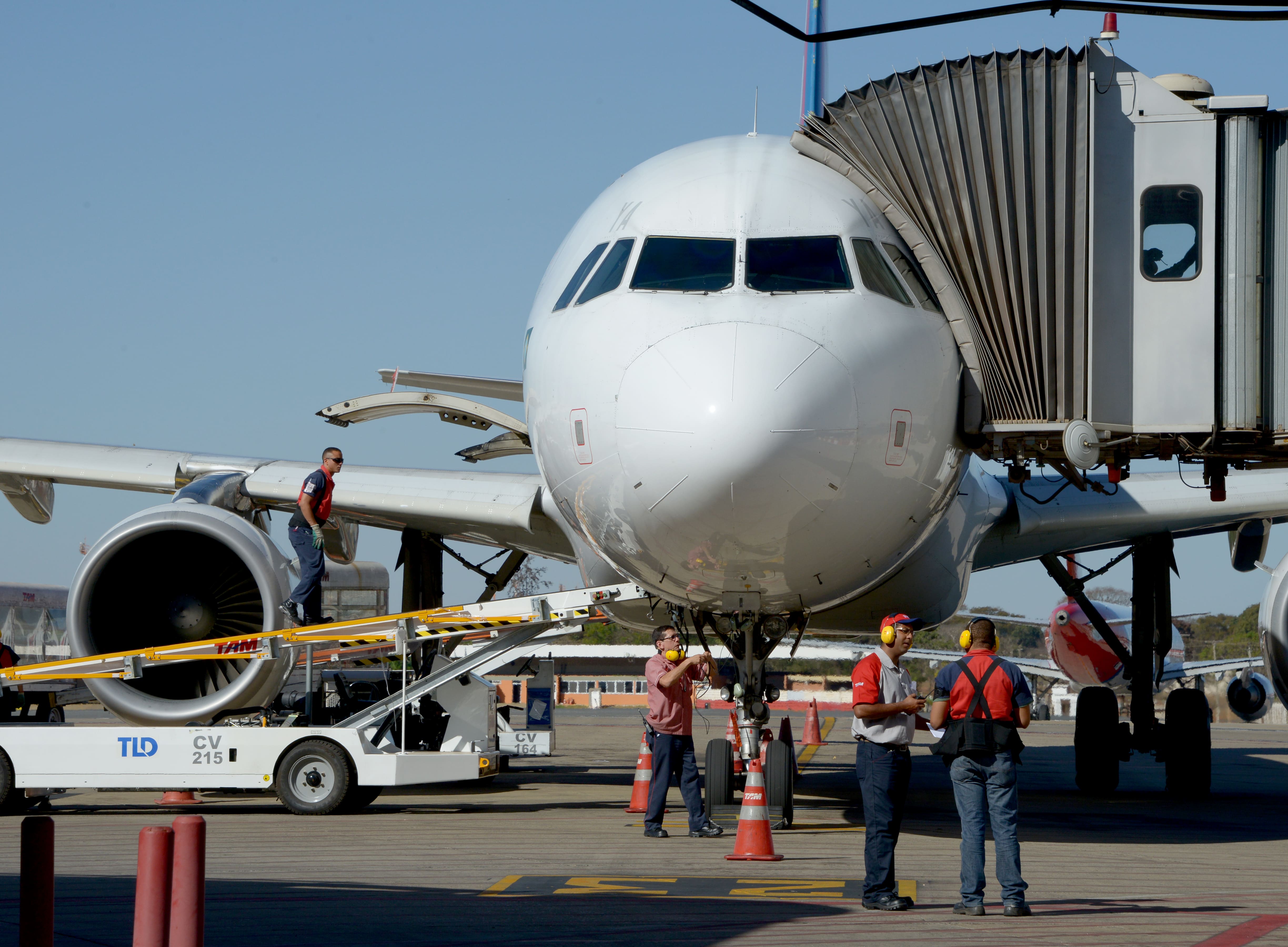 Foto mostra aeronave estacionada no finger com trabalhadores realizando inspeção e carregamento de carga.
