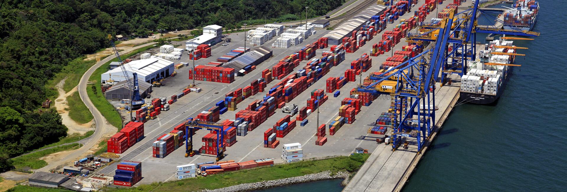 Vista aérea do Porto de Itaguaí, mostrando um terminal de contêineres com pilhas de contêineres, guindastes portuários, um navio atracado no cais e áreas de vegetação ao redor do complexo portuário.