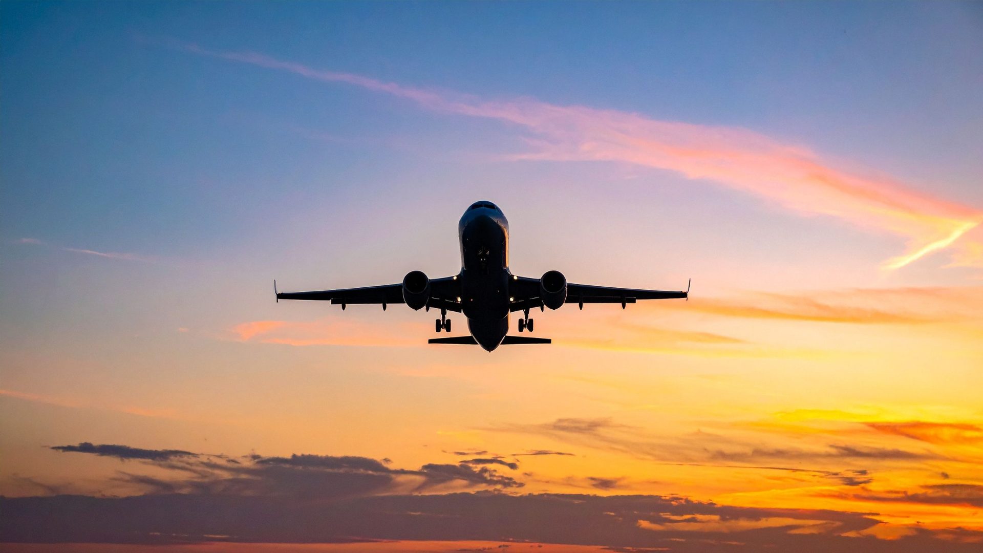 Fotografia de um avião comercial visto de frente e de baixo, em voo, com o trem de pouso acionado.