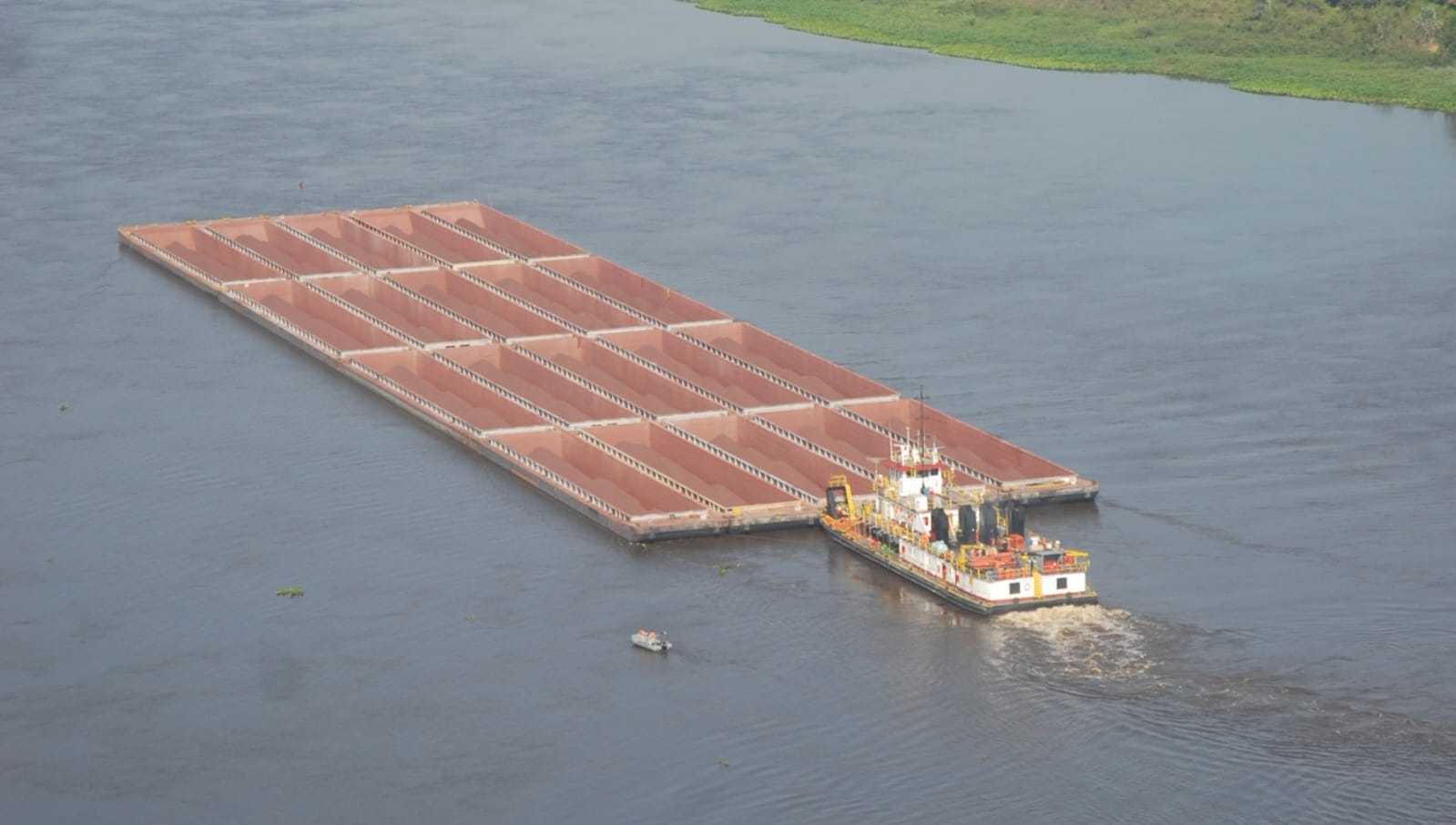 Foto aérea da Hidrovia do Paraguai mostrando um amplo trecho do rio de águas escuras e calmas. Em primeiro plano, um empurrador conduz um comboio formado por várias barcaças retangulares vazias, organizadas em fileiras. Ao redor, há extensas áreas de vegetação às margens e pequenas ilhas cobertas por gramíneas e arbustos, características do Pantanal. A paisagem se estende até o horizonte, envolta por uma leve névoa, evidenciando a dimensão da hidrovia e do ambiente natural.