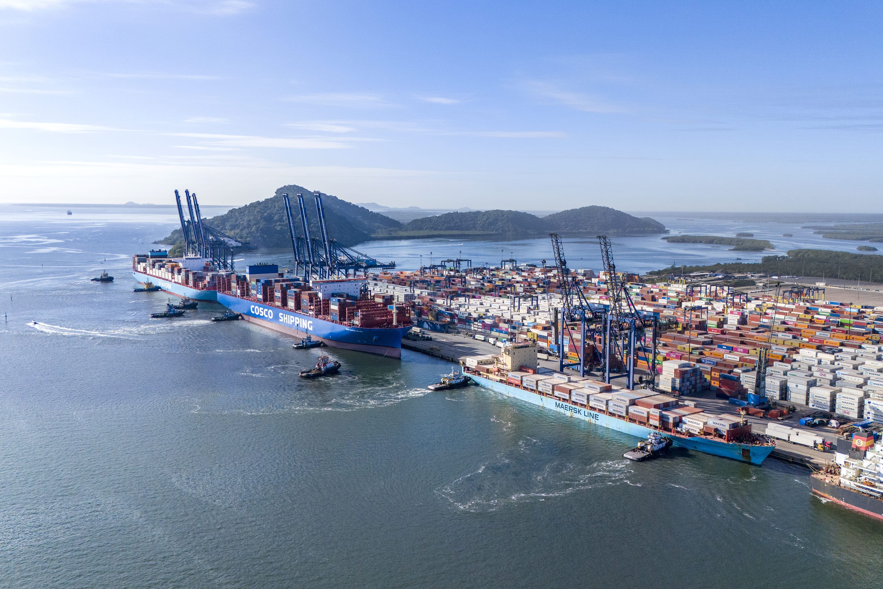 Imagem aérea de um terminal portuário de contêineres. Dois navios estão atracados junto ao cais, cercados por rebocadores. A área operacional reúne grandes pilhas de contêineres coloridos, guindastes portuários e equipamentos de movimentação de carga. Ao fundo, aparecem o mar, áreas de vegetação, ilhas e céu aberto.