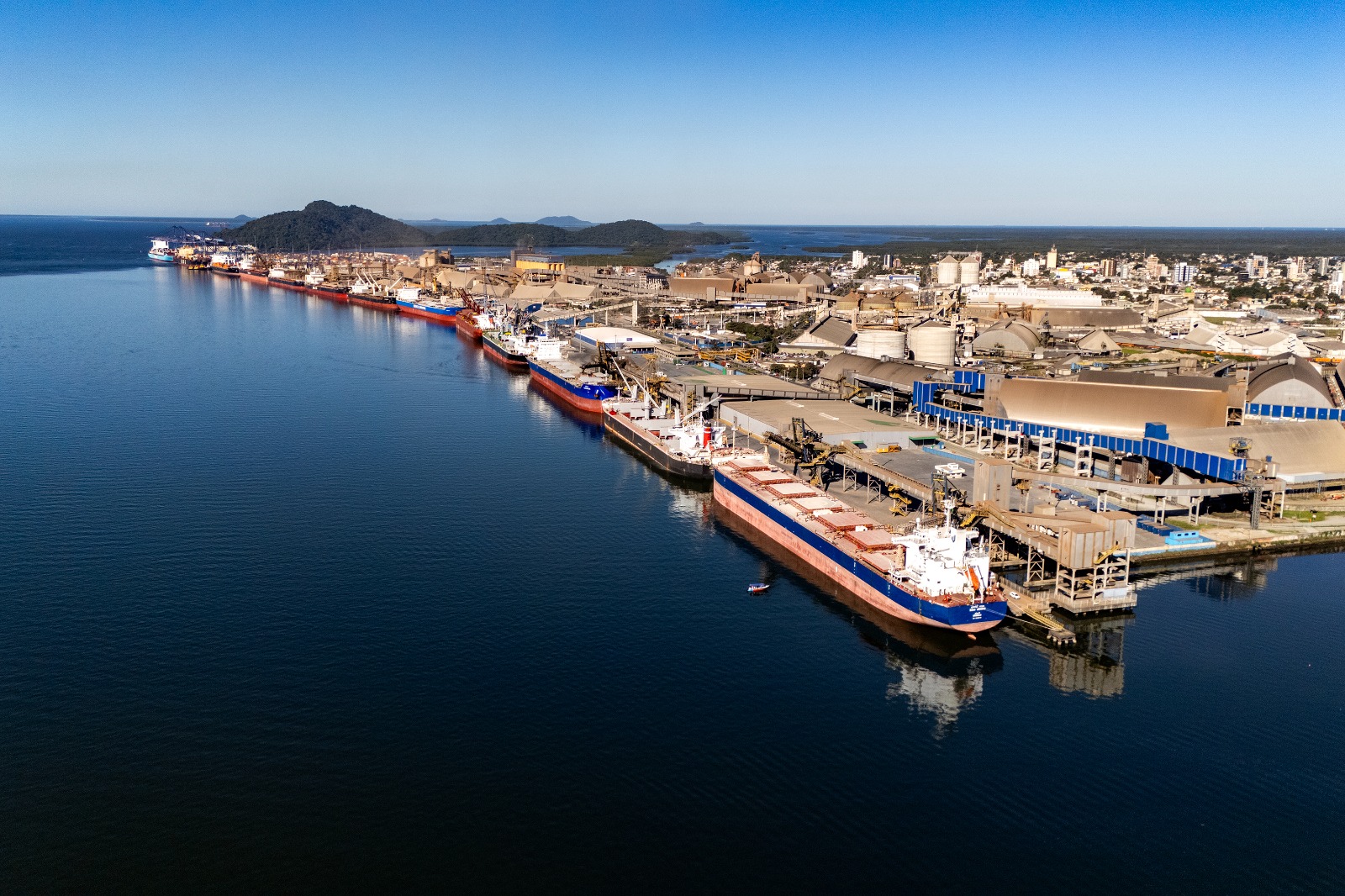 Imagem aérea de uma área portuária com navios atracados ao longo do cais. A foto mostra estruturas industriais, armazéns, silos, equipamentos de movimentação de carga e áreas operacionais próximas à água. Ao fundo, aparecem a cidade, áreas de vegetação, morros, ilhas e o mar sob céu aberto.