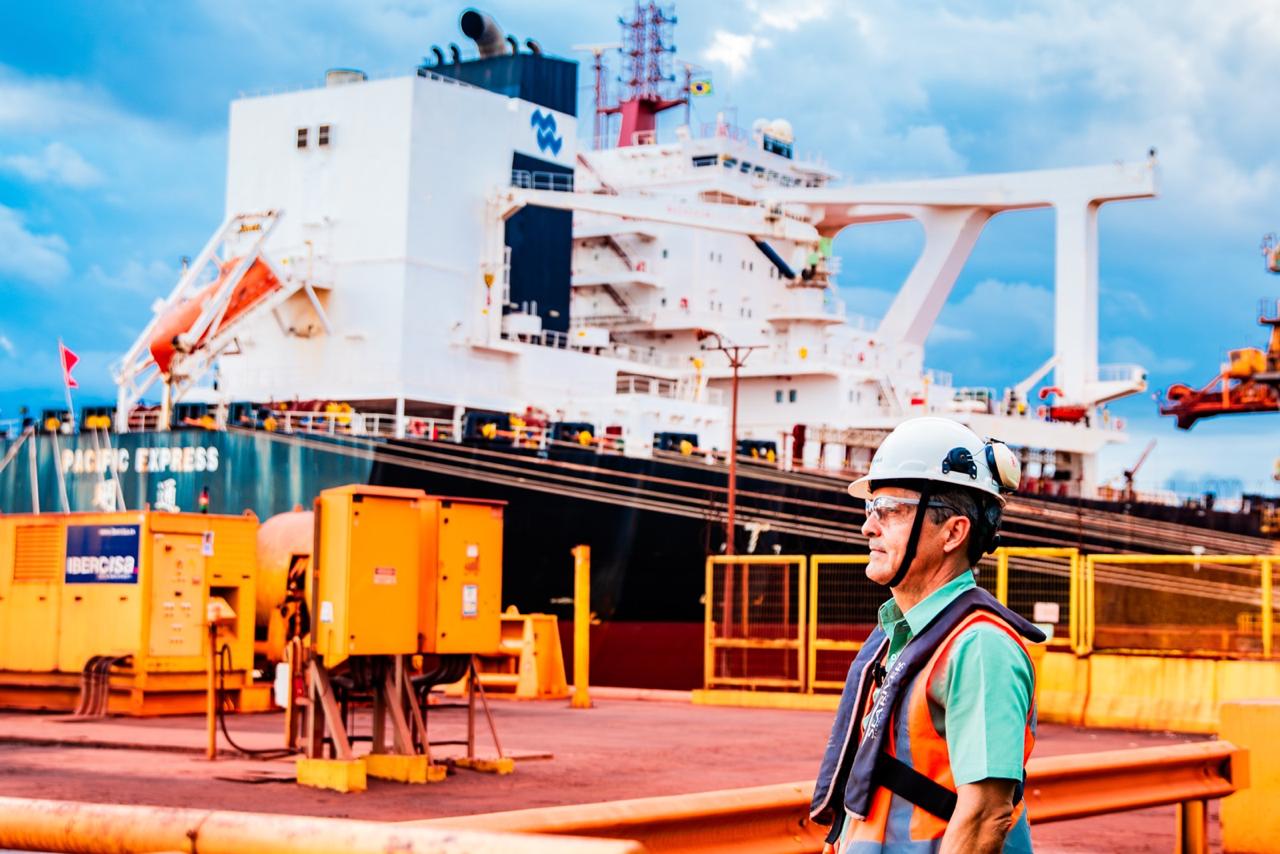 Imagem mostra um profissional em área portuária usando capacete, óculos de proteção e colete salva-vidas. Ao fundo, há um navio atracado, equipamentos operacionais, estruturas metálicas e áreas de circulação do terminal, sob céu parcialmente nublado.