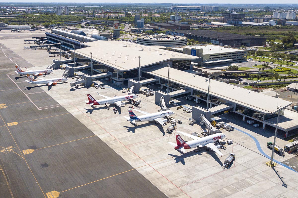 Vista aérea do Aeroporto Internacional Salgado Filho, em Porto Alegre (RS), mostrando o terminal de passageiros, o pátio de aeronaves e parte da pista. A imagem registra aviões de diferentes companhias aéreas estacionados junto às pontes de embarque, além da infraestrutura aeroportuária e da área urbana no entorno.