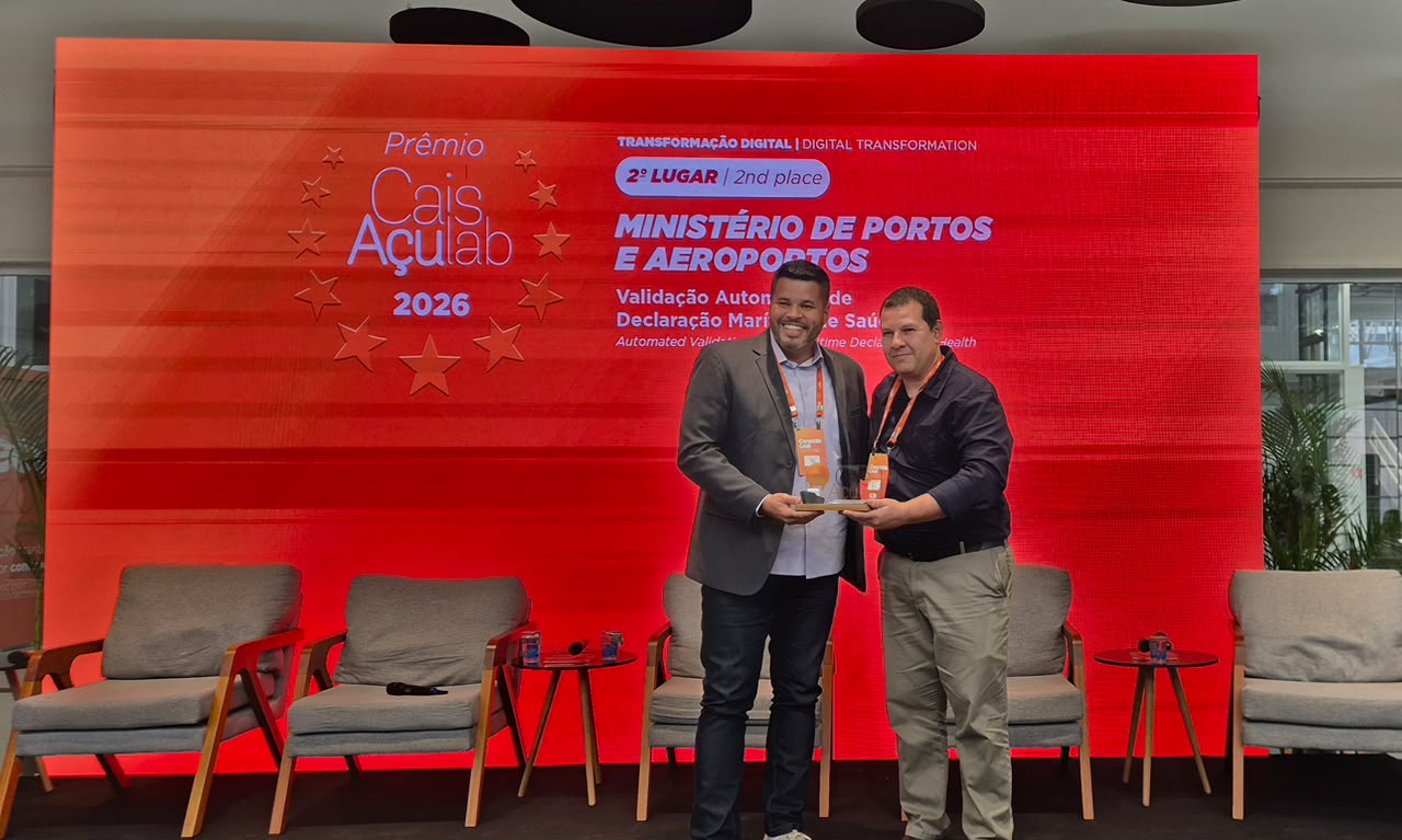 Dois representantes do Ministério de Portos e Aeroportos e do premio posam com o troféu do Prêmio Cais Açu Lab 2026, diante de painel que informa o 2º lugar na categoria Transformação Digital pelo projeto “Validação Automática de Declaração Marítima de Saúde”.