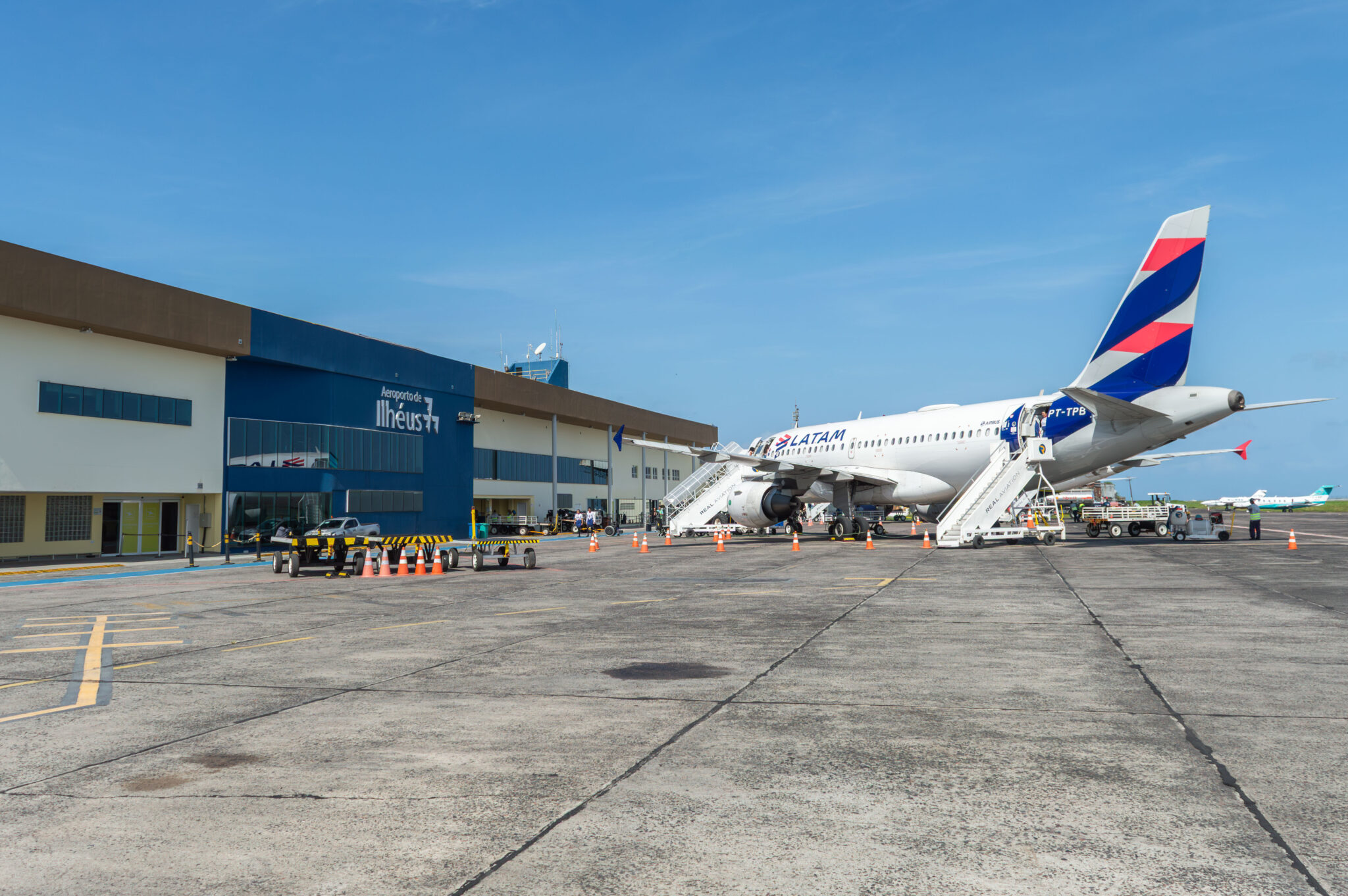 qual a descrição dessa imagem? Vista do pátio de aeronaves do Aeroporto Jorge Amado, em Ilhéus (BA), com avião comercial da LATAM estacionado em frente ao terminal de passageiros, escadas de embarque, equipamentos de apoio em solo e profissionais envolvidos nas operações aeroportuárias.