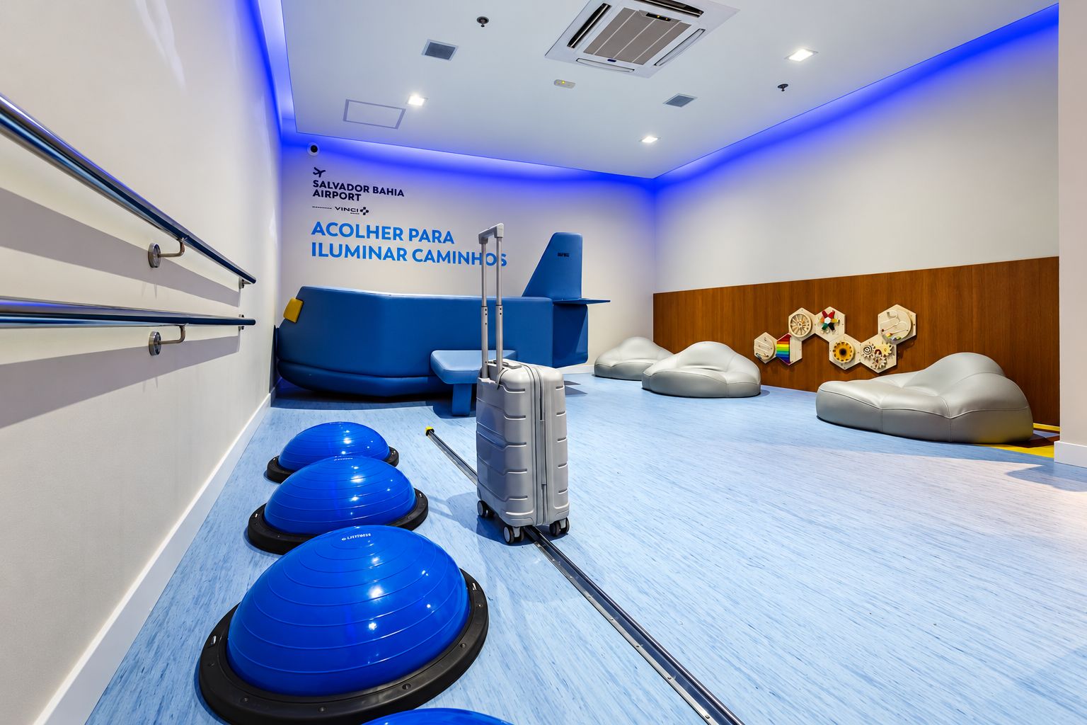 Sala com iluminação azul, sofá em formato de avião, mala, equipamentos de equilíbrio e pufes, no Aeroporto de Salvador.
