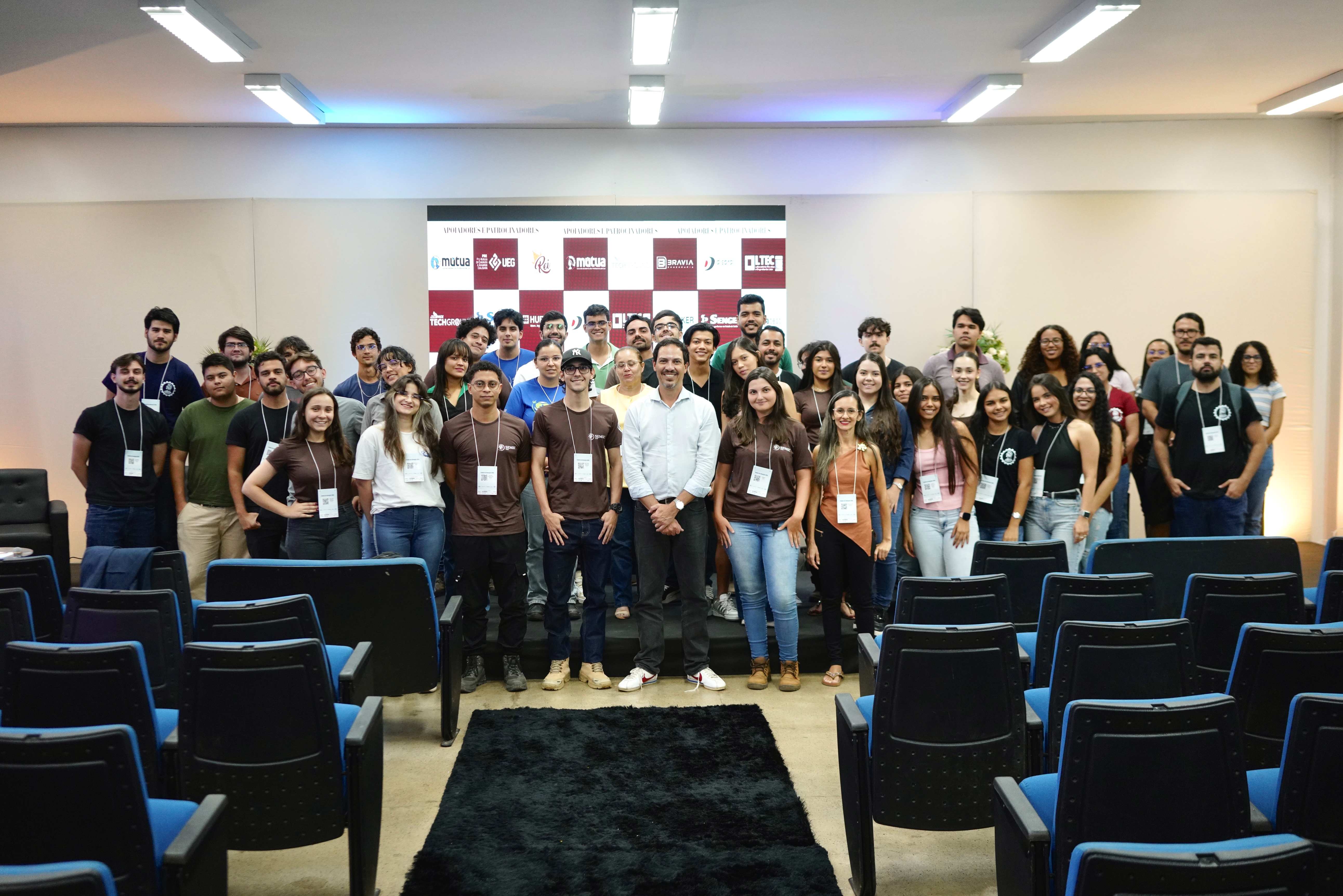 Grupo de participantes posa para foto em um auditório. Ao centro, um homem de camisa branca está cercado por jovens com crachás. À frente, aparecem fileiras de cadeiras azuis e pretas.