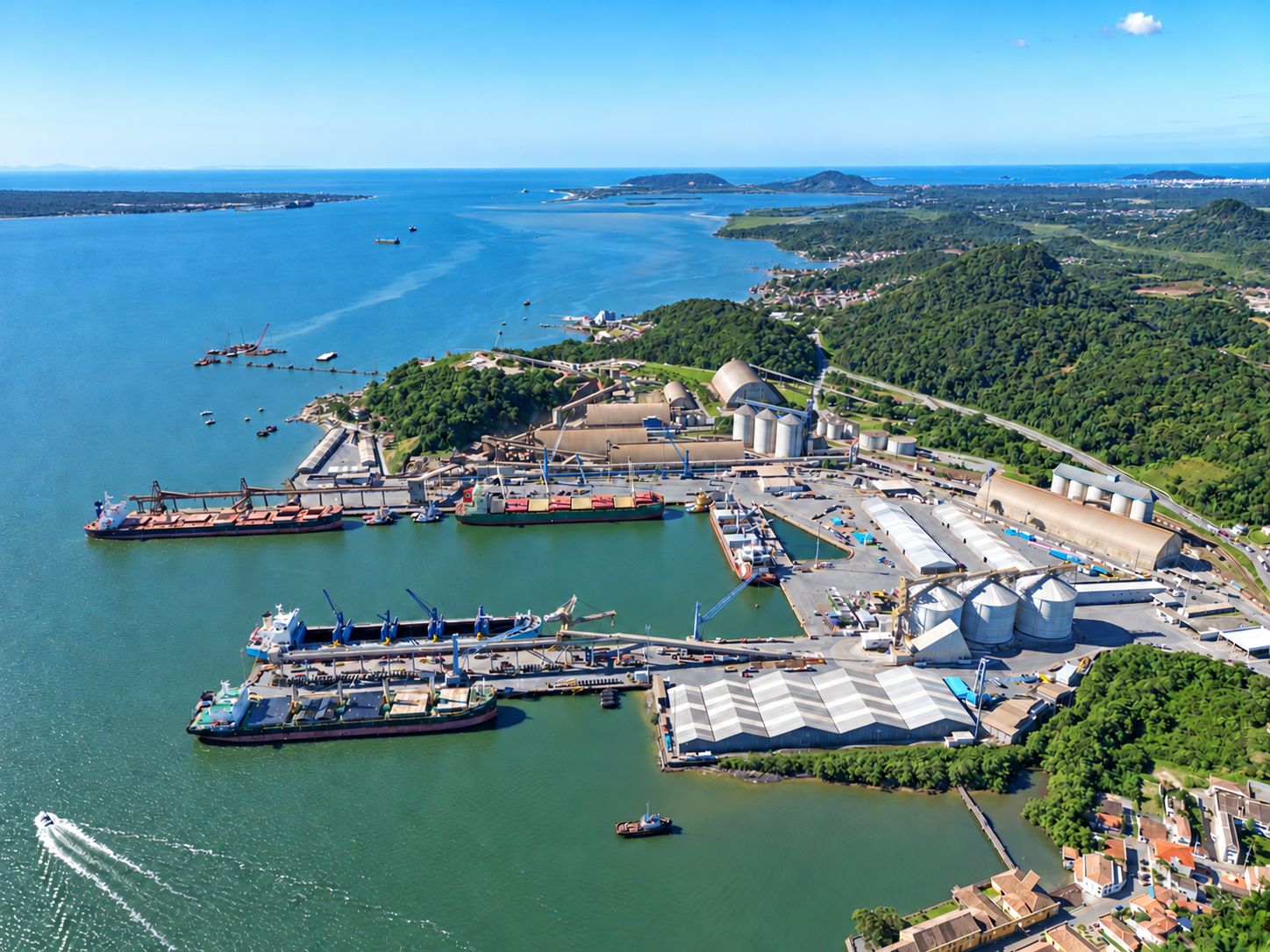 Vista aérea de da Baía de Babitonga (SC), com navios atracados, guindastes, armazéns e silos, cercado por água e áreas verdes.