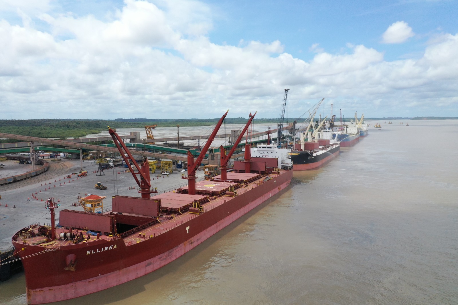 Vista aérea de um terminal portuário, com navios cargueiros atracados ao longo do cais para operações de movimentação de cargas. A imagem mostra guindastes, estruturas de transporte de granéis e instalações portuárias às margens de um rio, sob céu parcialmente nublado.