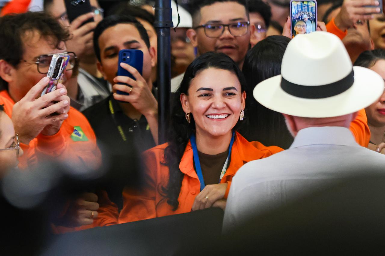 Público acompanha um evento atrás de uma grade de proteção. Em primeiro plano, uma mulher de cabelos escuros sorri enquanto segura um celular, cercada por outras pessoas que também observam a cerimônia e registram o momento com seus telefones. O ambiente é movimentado e transmite clima de expectativa e participação.