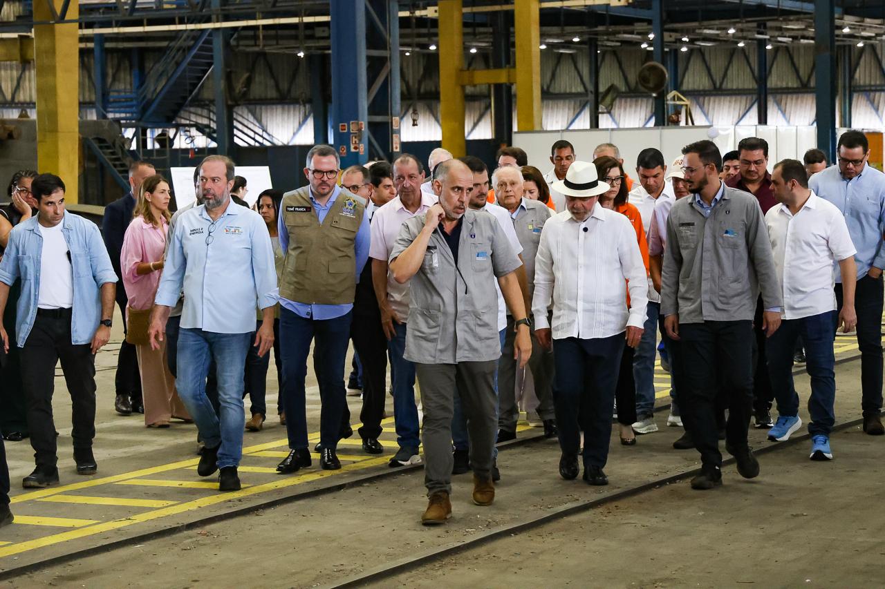A imagem registra uma comitiva de autoridades e lideranças políticas caminhando pelo interior de uma grande instalação industrial, com as características estruturais de uma fábrica ou estaleiro.