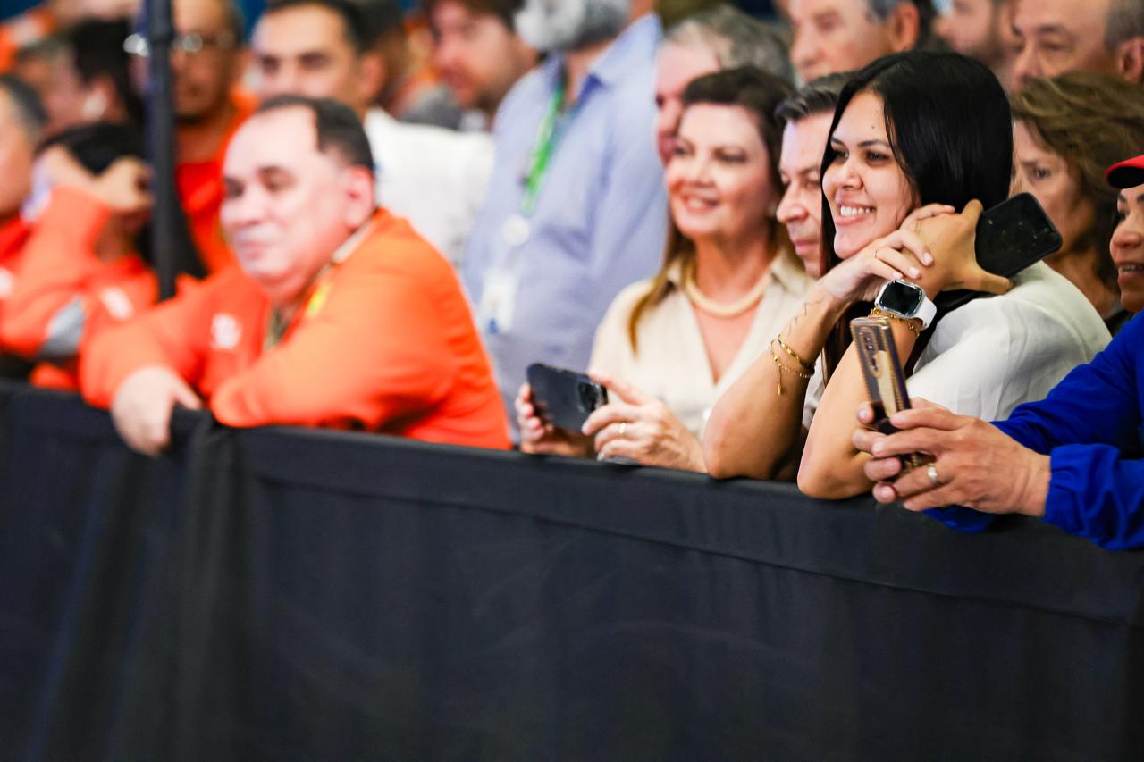 Público acompanha um evento atrás de uma grade de proteção. Em primeiro plano, uma mulher de cabelos escuros sorri enquanto segura um celular, cercada por outras pessoas que também observam a cerimônia e registram o momento com seus telefones. O ambiente é movimentado e transmite clima de expectativa e participação.