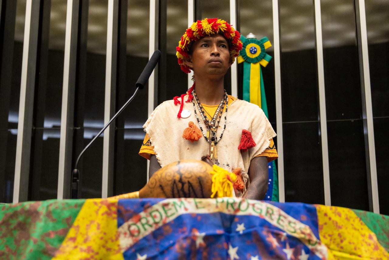Indígena posa para foto atrás de maracá em cima da bandeira nacional