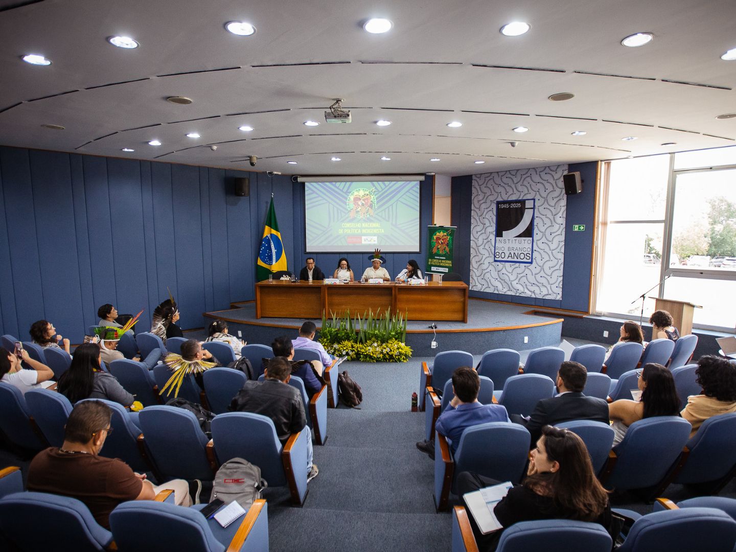 Reunião do Conselho Nacional de Política Indigenista (CNPI) em um auditório do Instituto Rio Branco. A imagem mostra uma mesa diretora com lideranças indígenas e autoridades, diante de uma plateia atenta que inclui representantes de povos originários e profissionais em trajes formais. O ambiente é de diálogo institucional e celebração dos 80 anos do Instituto.