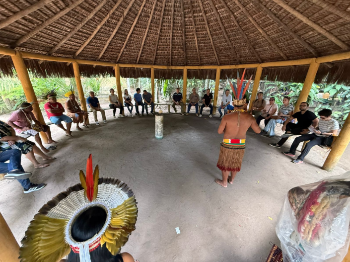 Indígena dialoga com pessoas em círculo embaixo de uma maloca