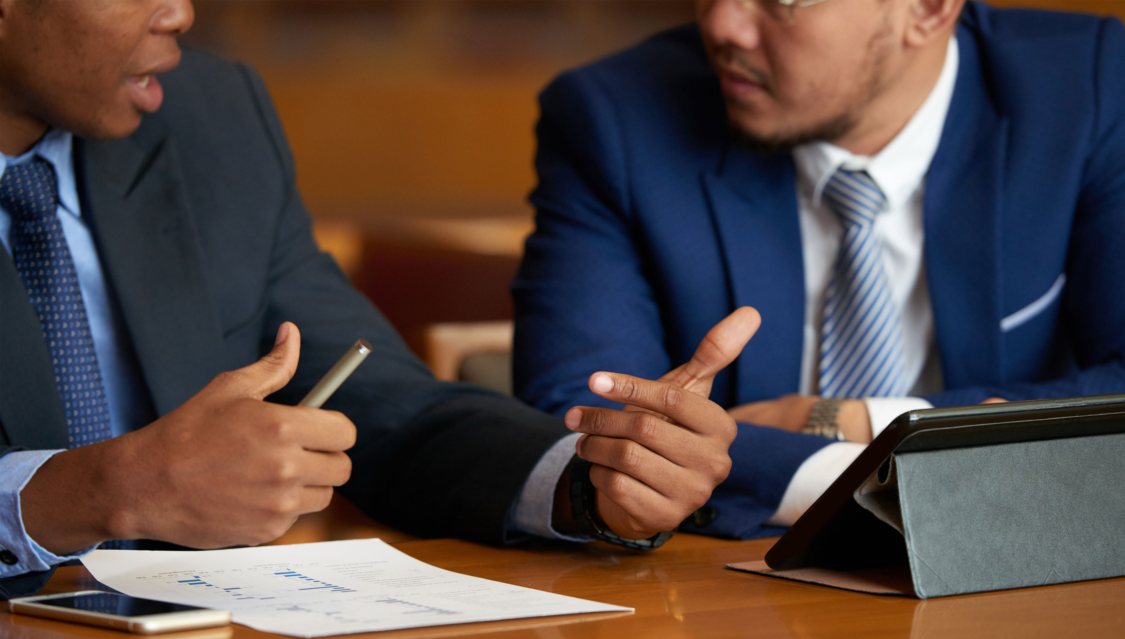Dois homens de paletó e gravata discutem sobre um tema, tendo um relatório e um tablet na mesa