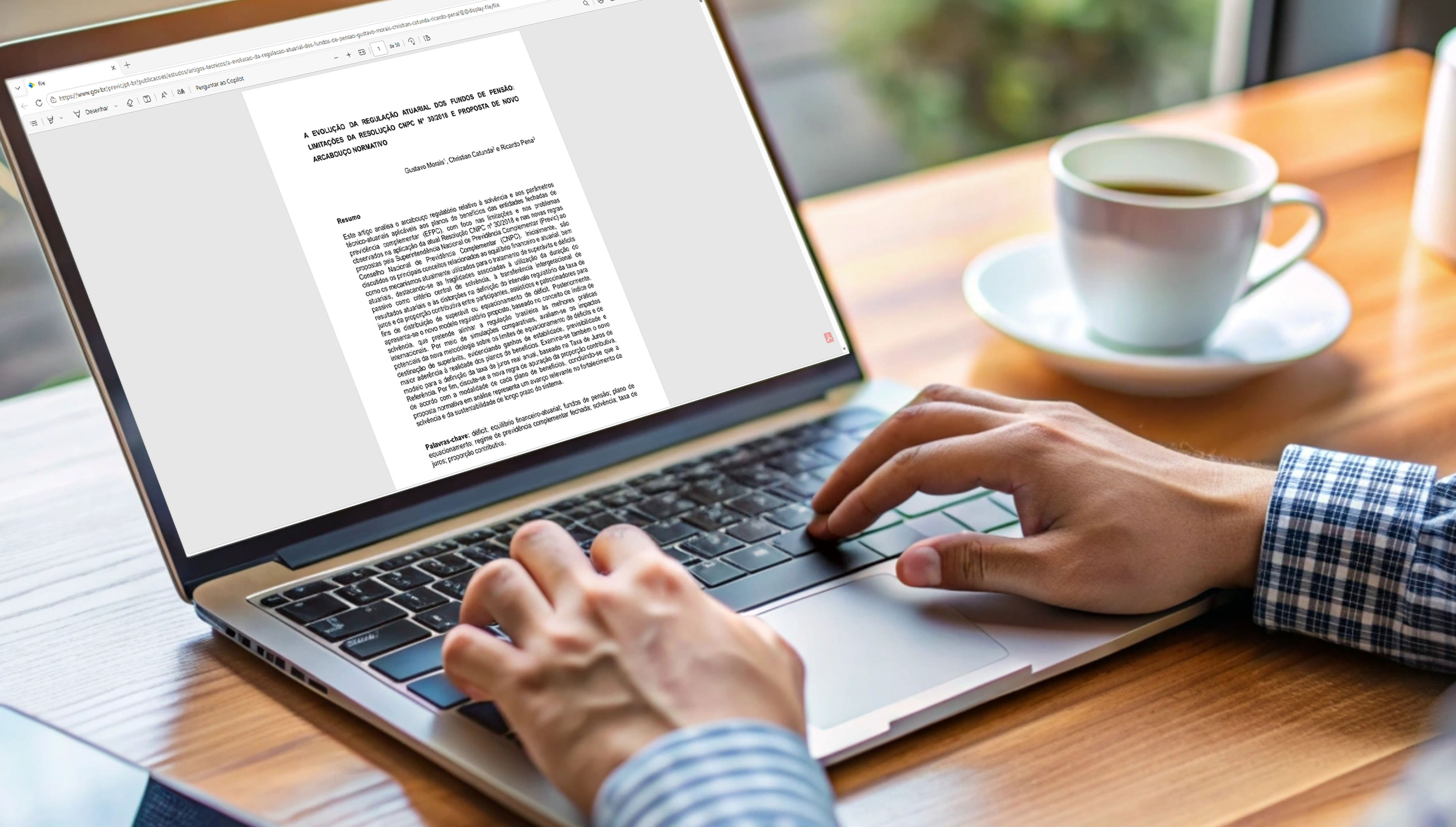 Pessoa digita artigo em computador sobre mesa de madeira, com xícara de café ao lado