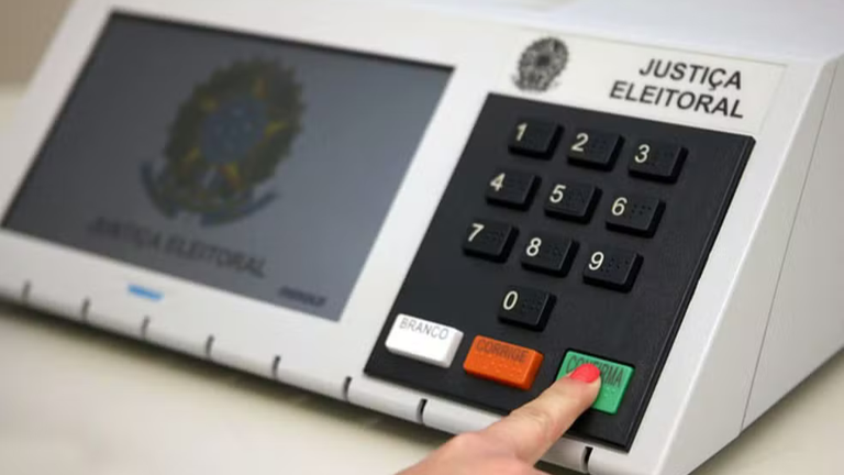 Urna eletrônica da Justiça Eleitoral em destaque, com uma pessoa pressionando a tecla verde “CONFIRMA” após digitar o voto..png