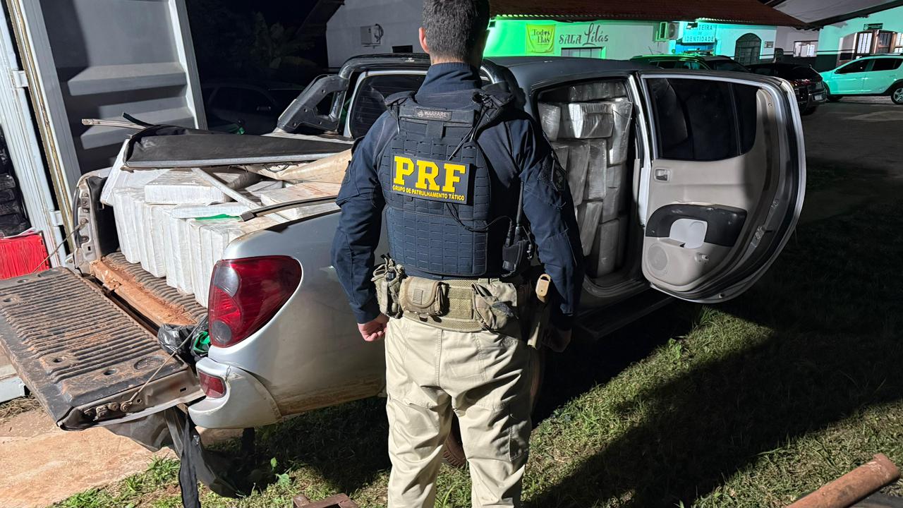 Uma imagem fotográfica colorida, em plano médio e capturada à noite, mostra um policial rodoviário federal de costas, em primeiro plano, diante da caçamba e da cabine aberta de uma picape carregada com fardos de droga.