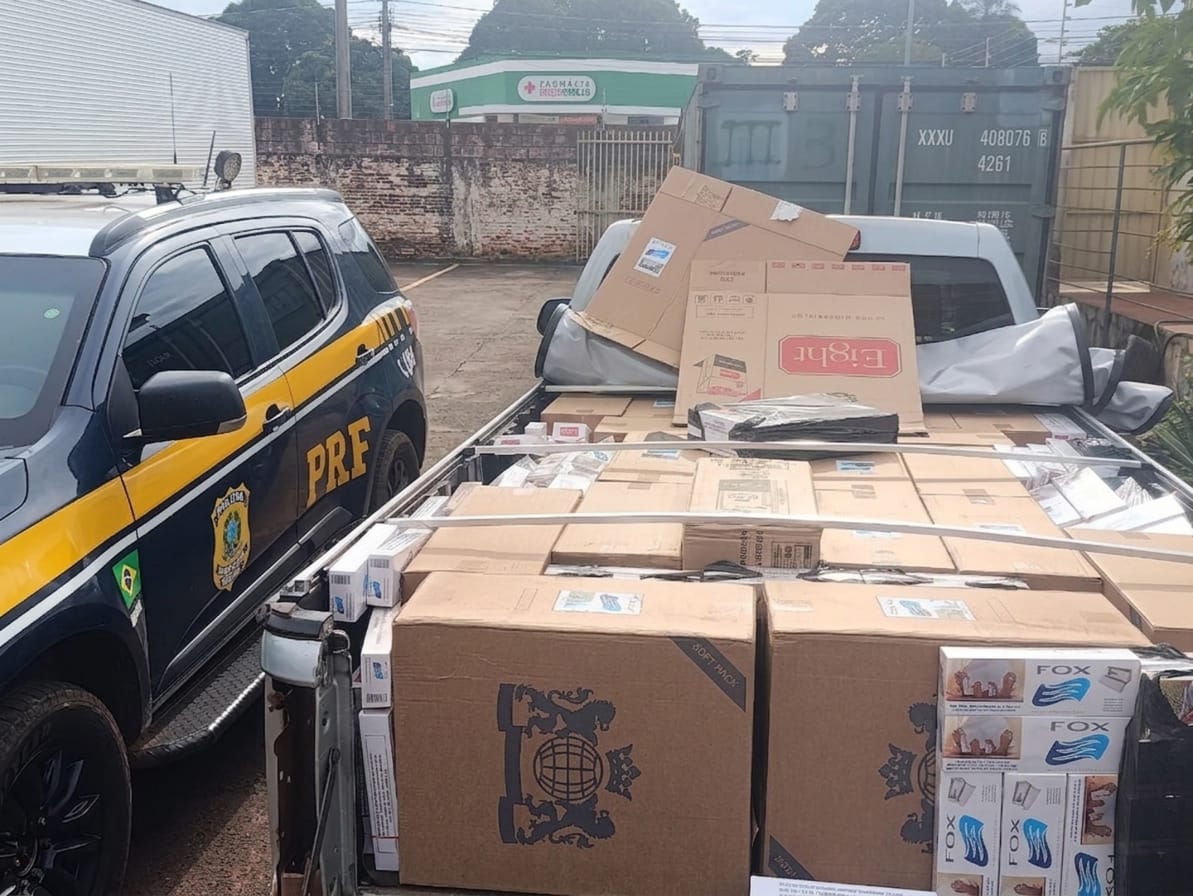 A foto mostra a caçamba de uma caminhonete carregada com caixas de contrabando, estacionada ao lado de uma viatura policial.  À esquerda: Vê-se a parte lateral dianteira de uma viatura da Polícia Rodoviária Federal (PRF), uma SUV de cor escura com os grafismos característicos em amarelo e o brasão da corporação na porta de motorista.  No centro/direita: A caçamba de uma caminhonete prata está completamente preenchida com grandes caixas de papelão pardo cheias de maços de cigarro. Algumas caixas na parte superior estão abertas e desfeitas, revelando marcas como "Eight" e "Fox".