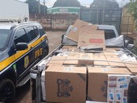 A foto mostra a caçamba de uma caminhonete carregada com caixas de contrabando, estacionada ao lado de uma viatura policial.  À esquerda: Vê-se a parte lateral dianteira de uma viatura da Polícia Rodoviária Federal (PRF), uma SUV de cor escura com os grafismos característicos em amarelo e o brasão da corporação na porta de motorista.  No centro/direita: A caçamba de uma caminhonete prata está completamente preenchida com grandes caixas de papelão pardo cheias de maços de cigarro. Algumas caixas na parte superior estão abertas e desfeitas, revelando marcas como "Eight" e "Fox".