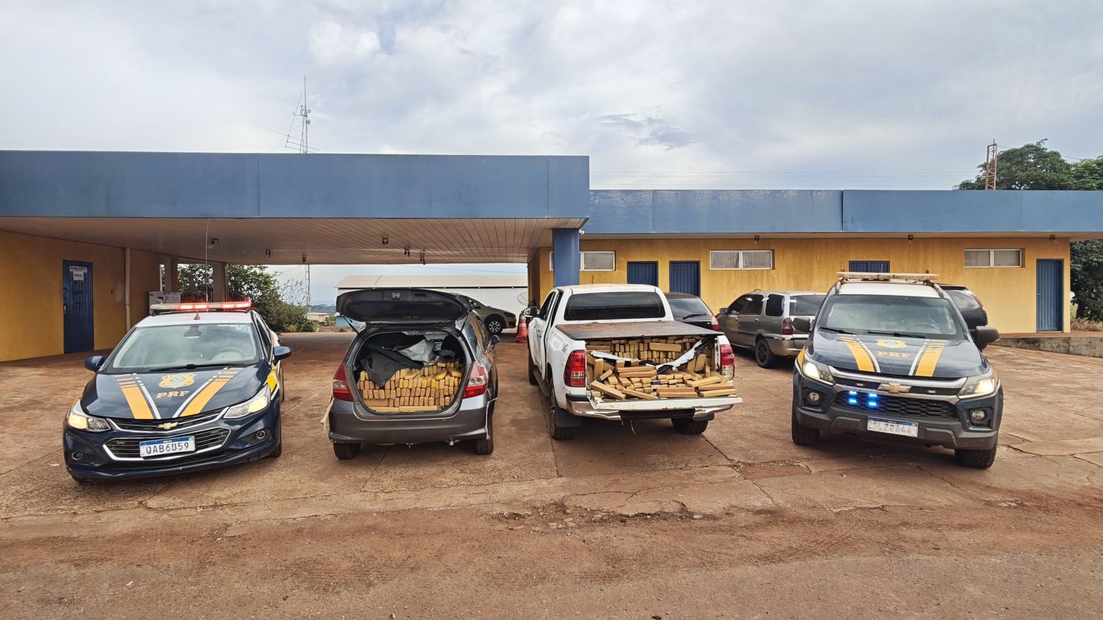A imagem mostra uma apreensão de drogas realizada pela Polícia Rodoviária Federal (PRF) no Brasil.  Em frente a um posto policial de cor amarela e azul, estão estacionados quatro veículos alinhados lado a lado:  Nas extremidades: Duas viaturas oficiais da PRF (uma caminhonete Chevrolet S10 à direita e um carro de passeio Chevrolet Cruze à esquerda) com os girofles e luzes intermitentes acesos.  No centro: Dois veículos civis apreendidos — um carro de passeio cinza com o porta-malas aberto e uma caminhonete branca com a caçamba aberta.  Ambos os veículos do centro estão completamente carregados com dezenas de tabletes prensados de maconha (de cor marrom/amarelada), empilhados até o limite do espaço interno e da caçamba.
