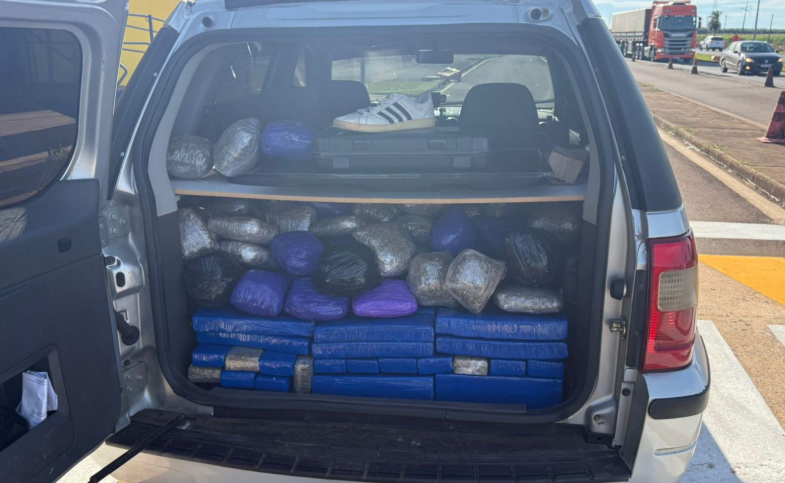 A imagem mostra o porta-malas aberto de um veículo utilitário desportivo (SUV) de cor prata, completamente carregado com fardos e pacotes de drogas.