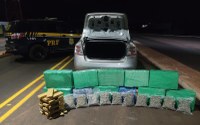 A imagem mostra uma apreensão de drogas realizada pela Polícia Rodoviária Federal (PRF) à noite, em uma rodovia.  Em primeiro plano, no chão, há uma grande quantidade de entorpecentes apreendidos organizados em fileiras:  Vários fardos grandes retangulares envoltos em plástico verde.  Uma fileira de sacos plásticos transparentes contendo uma substância esverdeada em pedaços (skunk/botões de maconha).  Uma pilha menor de tabletes marrons (prensados) no canto esquerdo.  Logo atrás das drogas, está a traseira de um carro de passeio prata com o porta-malas aberto e vazio. À esquerda, estacionada na pista ao lado de faixas amarelas duplas, está uma viatura preta e amarela da PRF. O cenário de fundo é escuro, indicando um ambiente de estrada durante a noite.