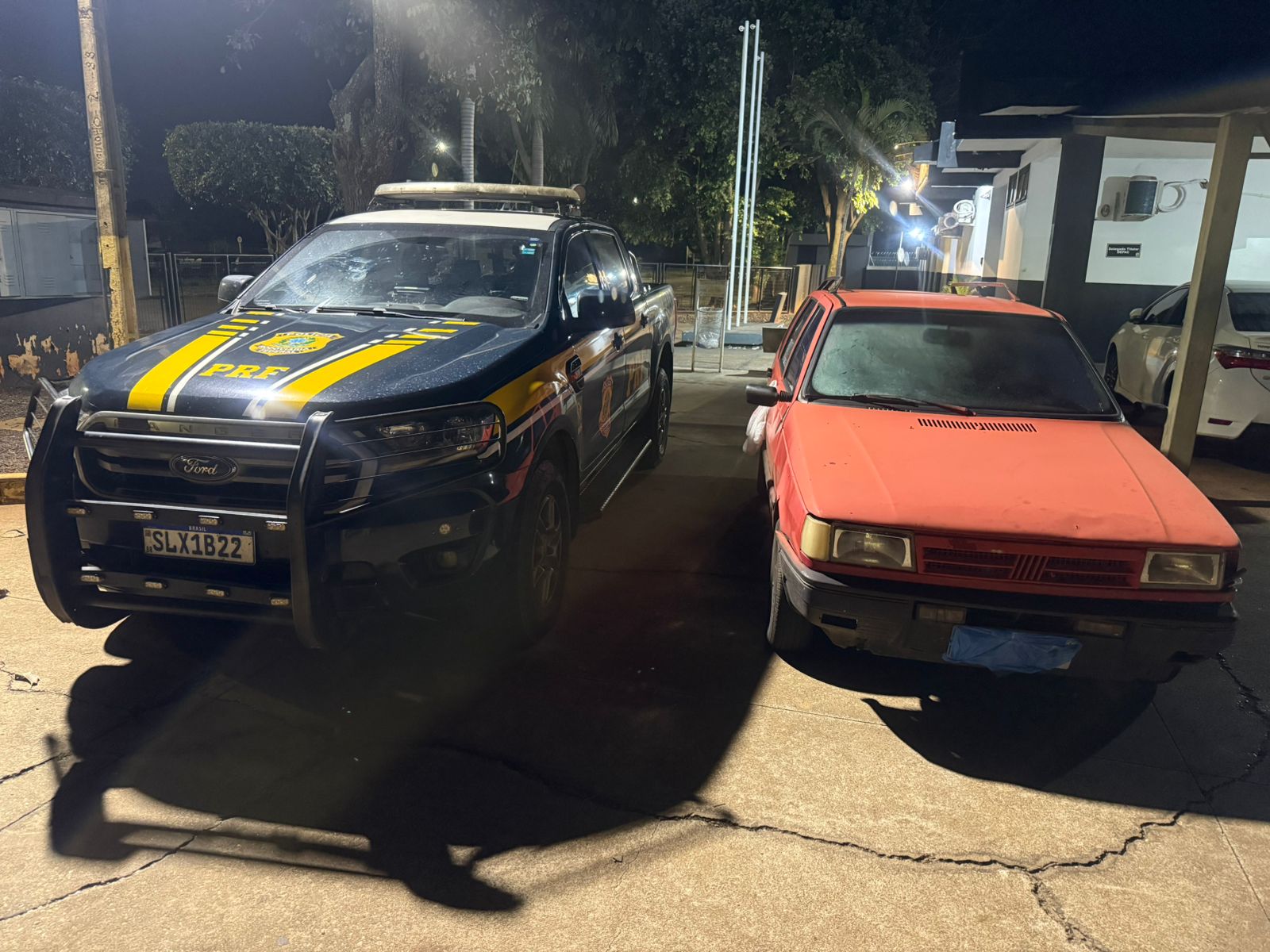 A imagem mostra dois carros, uma viatura da PRF e um Fiat/Elba vermelho, estacionados lado a lado à noite