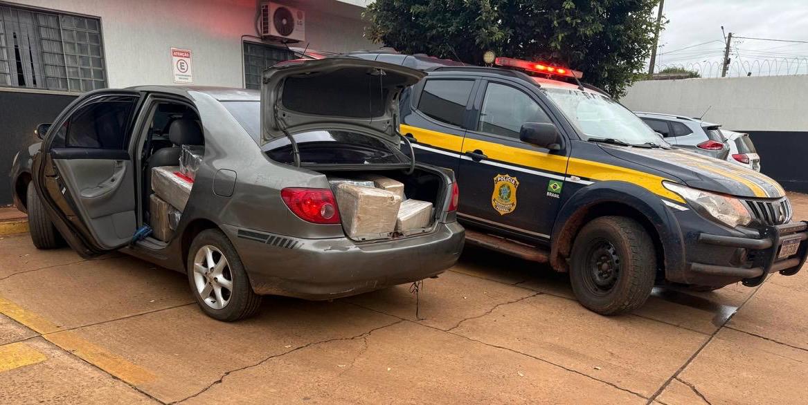 Uma viatura da Polícia Rodoviária Federal (PRF) estacionada ao lado de um veículo de passeio com o porta-malas e as portas traseiras abertas, revelando fardos de droga em seu interior. A imagem está em plano médio e horizontal.