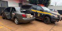 Uma viatura da Polícia Rodoviária Federal (PRF) estacionada ao lado de um veículo de passeio com o porta-malas e as portas traseiras abertas, revelando fardos de droga em seu interior. A imagem está em plano médio e horizontal.
