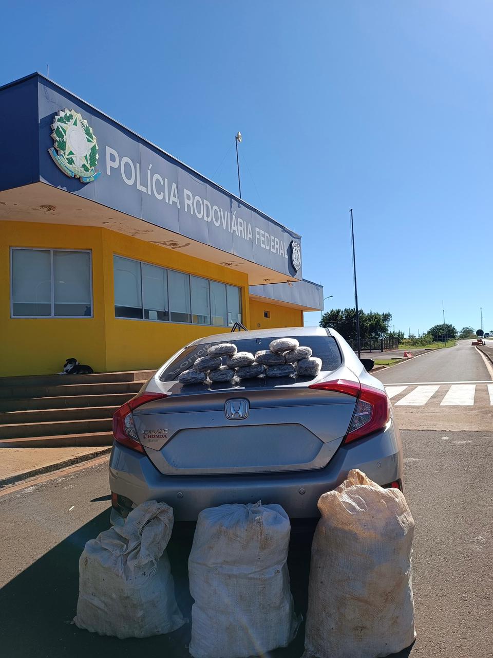 O local: Um posto da Polícia Rodoviária Federal (PRF), com uma fachada amarela e azul clara sob um céu limpo.  O veículo: Um carro modelo Honda Civic de cor prata, estacionado de costas em frente ao posto.  A apreensão: Vários tabletes embalados estão empilhados em cima do porta-malas do veículo, e três grandes sacos de Linhagem/estopa brancos estão posicionados no chão, logo atrás do carro.