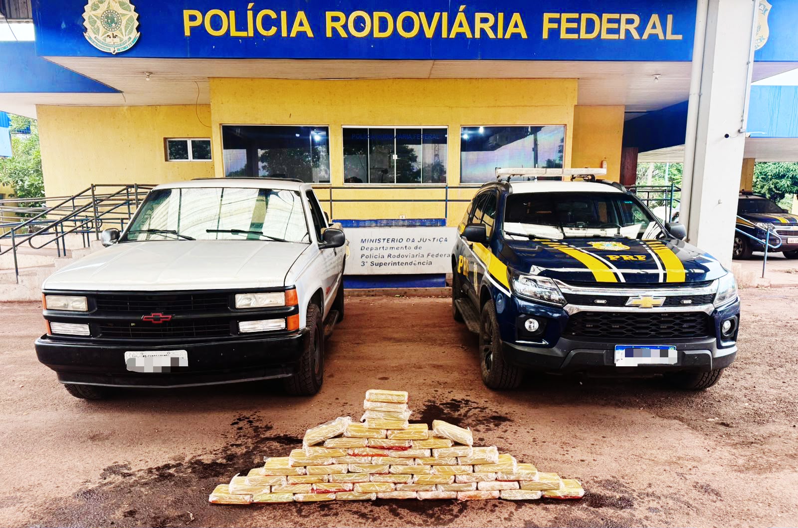 Aqui estão os elementos principais da foto de forma simples:  Veículos: Dois veículos estão estacionados lado a lado. À esquerda, há uma caminhonete branca mais antiga (Chevrolet). À direita, uma viatura oficial da PRF, de cor azul escura com faixas amarelas.  Apreensão: No chão, logo à frente dos veículos, há uma grande quantidade de tabletes de droga (cocaína) empilhados em formato de pirâmide.