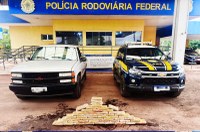 Aqui estão os elementos principais da foto de forma simples:  Veículos: Dois veículos estão estacionados lado a lado. À esquerda, há uma caminhonete branca mais antiga (Chevrolet). À direita, uma viatura oficial da PRF, de cor azul escura com faixas amarelas.  Apreensão: No chão, logo à frente dos veículos, há uma grande quantidade de tabletes de droga (cocaína) empilhados em formato de pirâmide.