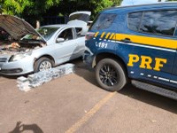Em primeiro plano, aparece a lateral traseira de uma viatura oficial da Polícia Rodoviária Federal (PRF), na cor azul com faixas amarelas, exibindo a sigla "PRF" e o número de emergência "191".  Lado esquerdo: Um carro de passeio prata (modelo Volkswagen) está estacionado com o capô e o porta-malas totalmente abertos.  Ao centro: No chão, logo à frente do pneu dianteiro do carro prata, há dezenas de tabletes de entorpecentes embalados e organizados em fileiras.