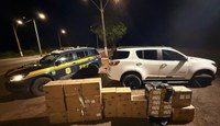 À esquerda, está uma viatura oficial da Polícia Rodoviária Federal (PRF), modelo Chevrolet Trailblazer. O veículo é azul-escuro com detalhes amarelos, incluindo a inscrição "PRF" em destaque na lateral traseira e o brasão da corporação na porta dianteira. Os faróis do carro estão acesos.  À direita, está um SUV de cor branca, também modelo Chevrolet Trailblazer, que parece ser o veículo abordado na ocorrência.  Em primeiro plano, no chão de terra à frente dos dois veículos, há uma grande quantidade de mercadorias apreendidas.