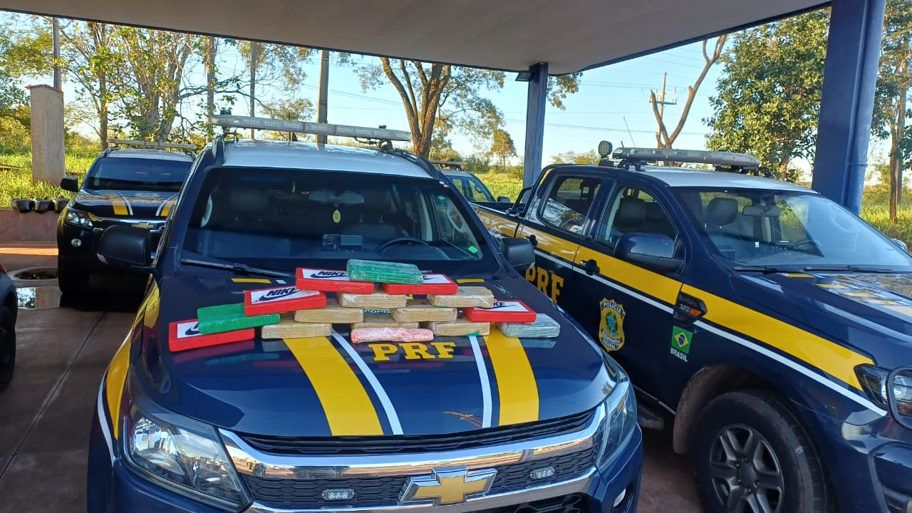 Imagem frontal de viaturas da Polícia Rodoviária Federal (PRF) estacionadas sob uma cobertura, com materiais apreendidos em destaque. Em primeiro plano: Vários tabletes de drogas (embalados em cores como marrom, vermelho com logotipos e verde) estão empilhados sobre o capô de uma viatura Chevrolet azul e amarela.