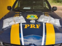 A imagem mostra a capô de uma viatura da Polícia Rodoviária Federal (PRF) do Brasil, com as cores características azul e amarela e o brasão da instituição no centro.  Sobre o capô do veículo, estão dispostos os materiais apreendidos em uma operação:  Quatro pacotes plásticos grandes transparentes, contendo diversos tabletes menores embalados (provavelmente medicamentos ou entorpecentes).  Um tablete menor e isolado, envolto em fita ou plástico de cor cinza/clara, posicionado bem no centro, logo abaixo do brasão da PRF.