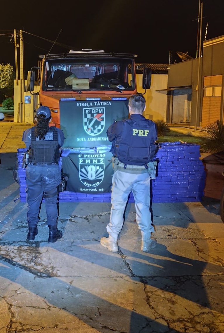 Dois policiais, vistos de costas, À frente deles, dezenas de tabletes retangulares embalados em plástico azul escuro estão empilhados em formato de muro, cobrindo a parte inferior do caminhão. estão em pé no centro da imagem, posicionados diante de um caminhão de cor laranja e de uma grande quantidade de tabletes de droga apreendida..