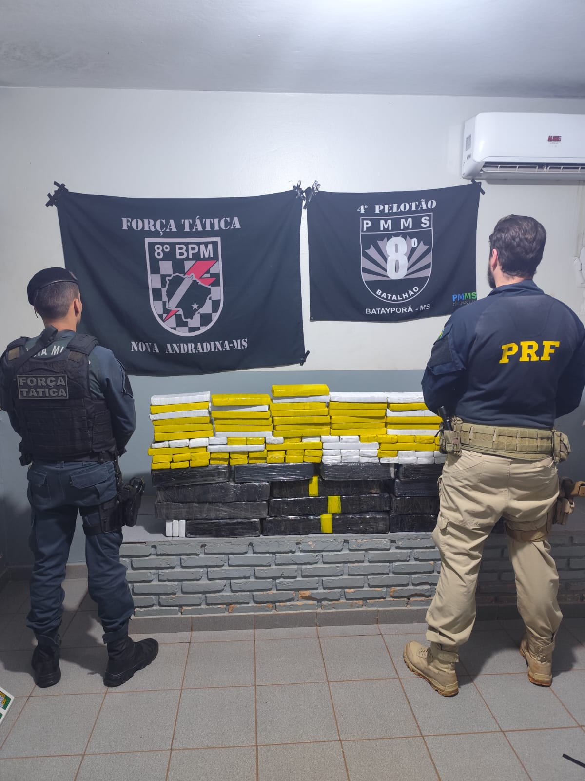 Dois policiais aparecem de costas observando uma grande pilha de tabletes de entorpecentes empilhados contra uma mureta de tijolos cinza.  Policiais: À esquerda, um policial militar da Força Tática (8º BPM - Nova Andradina-MS) usa fardamento operacional escuro. À direita, um policial rodoviário federal (PRF) veste calça caqui e jaqueta azul-escura com a sigla da corporação em amarelo.