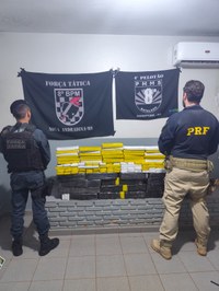 Dois policiais aparecem de costas observando uma grande pilha de tabletes de entorpecentes empilhados contra uma mureta de tijolos cinza.  Policiais: À esquerda, um policial militar da Força Tática (8º BPM - Nova Andradina-MS) usa fardamento operacional escuro. À direita, um policial rodoviário federal (PRF) veste calça caqui e jaqueta azul-escura com a sigla da corporação em amarelo.