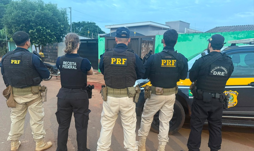 Foto tirada de costas mostrando cinco agentes de segurança pública brasileiros em uma rua, observando uma casa ao fundo. Eles estão parados lado a lado, vestindo coletes balísticos com as identificações de suas respectivas instituições. Da esquerda para a direita: Três agentes da PRF (Polícia Rodoviária Federal): Vestem coletes pretos com a sigla PRF em letras amarelas e calças táticas bege-claras. O quarto agente possui uma tarjeta abaixo da sigla escrita "Núcleo de Operações Especiais". Uma agente da Receita Federal (segunda da esquerda): Veste colete preto com a inscrição RECEITA FEDERAL em letras brancas e calça tática preta. Um agente da Polícia Federal (último à direita): Veste colete e calça pretos, com a inscrição POLÍCIA FEDERAL / DRE (Delegacia de Repressão a Entorpecentes) em letras brancas.