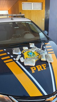 Viatura: Um veículo preto com grafismos em amarelo e branco. No capô, destaca-se o brasão oficial da corporação com a inscrição "POLÍCIA RODOVIÁRIA FEDERAL" e a sigla "PRF" logo abaixo.  Apreensão: Sobre o capô, há vários tabletes e pacotes pequenos envoltos em plástico transparente, contendo substâncias ilícitas apreendidas.