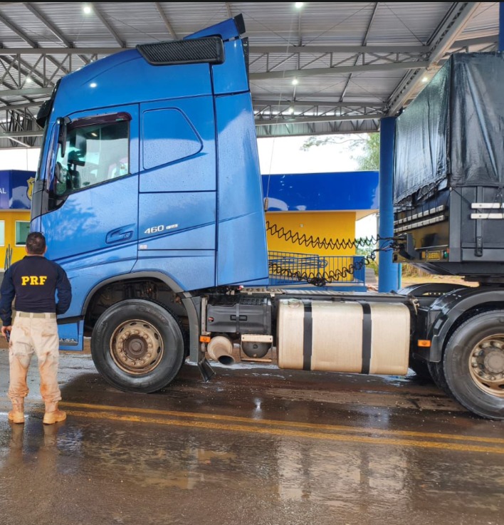 A imagem mostra um caminhão de grande porte azul estacionado de perfil sob a cobertura de um posto ou unidade operacional.  À esquerda: Um policial rodoviário federal (identificado pela inscrição "PRF" nas costas da farda escura) aparece de costas, observando o veículo. Ele veste calça clara e botas.  Ao fundo: É possível ver uma estrutura de apoio com as cores azul e amarela, típicas de postos da Polícia Rodoviária Federal (PRF).