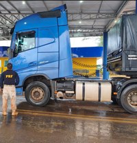 A imagem mostra um caminhão de grande porte azul estacionado de perfil sob a cobertura de um posto ou unidade operacional.  À esquerda: Um policial rodoviário federal (identificado pela inscrição "PRF" nas costas da farda escura) aparece de costas, observando o veículo. Ele veste calça clara e botas.  Ao fundo: É possível ver uma estrutura de apoio com as cores azul e amarela, típicas de postos da Polícia Rodoviária Federal (PRF).
