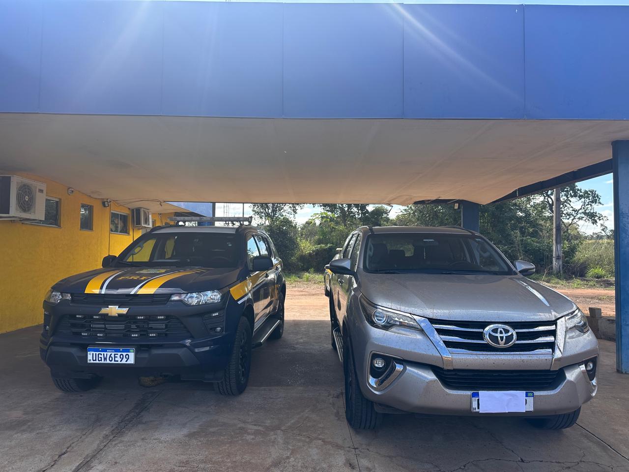 Imagem frontal de dois carros estacionados lado a lado sob uma cobertura.  À esquerda: Uma viatura azul e amarela da Polícia Rodoviária Federal (PRF), modelo Chevrolet.  À direita: Uma caminhonete SUV prata, modelo Toyota SW4, com a placa tampada por um quadrado branco.