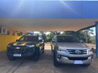 Imagem frontal de dois carros estacionados lado a lado sob uma cobertura.  À esquerda: Uma viatura azul e amarela da Polícia Rodoviária Federal (PRF), modelo Chevrolet.  À direita: Uma caminhonete SUV prata, modelo Toyota SW4, com a placa tampada por um quadrado branco.