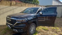 Uma caminhonete preta da marca Ram, modelo Rampage, estacionada em um terreno de terra e grama baixa, ao lado de um muro de concreto com cerca de arame farpado no topo.